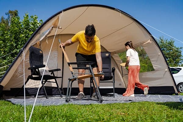 Amplia tienda de campaña tipo iglú con avance en un campo de hierba, dos personas preparando sillas y mesa de camping bajo un cielo azul soleado